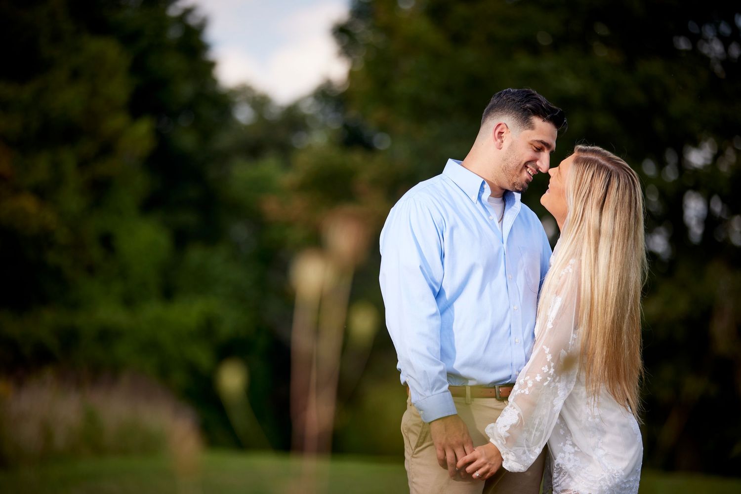 Couple sharing a romantic moment outdoors, man in blue shirt and woman in lace top touching foreheads in a garden.