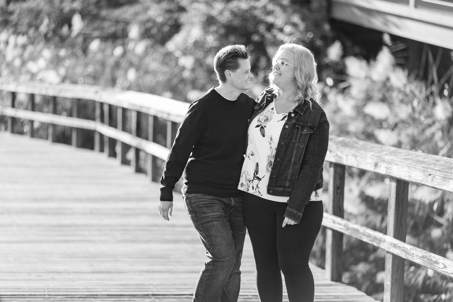 Black and white photo of a couple walking and kissing on a wooden boardwalk in a park.