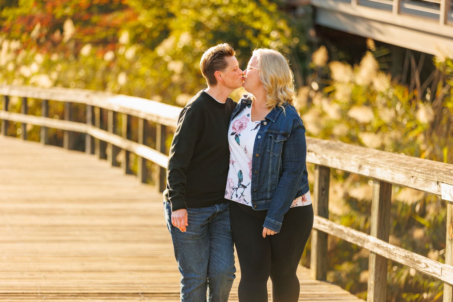 Couple sharing a sweet kiss on a sunny wooden boardwalk surrounded by warm autumn foliage.