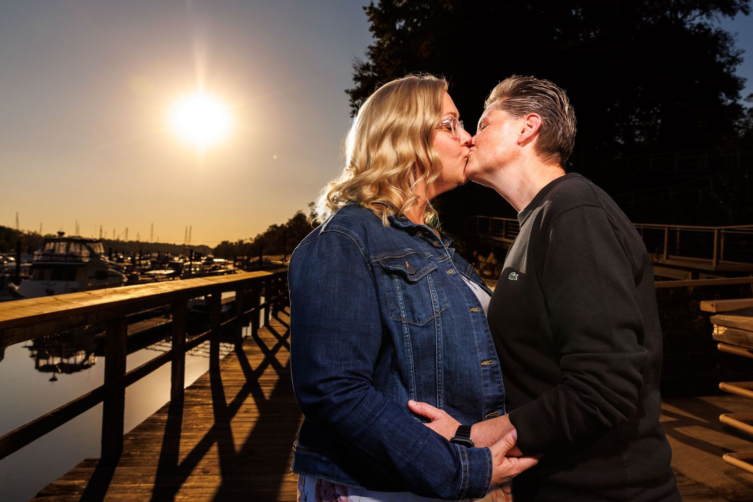 Couple sharing a romantic kiss on a waterfront dock at golden sunset with marina in background.