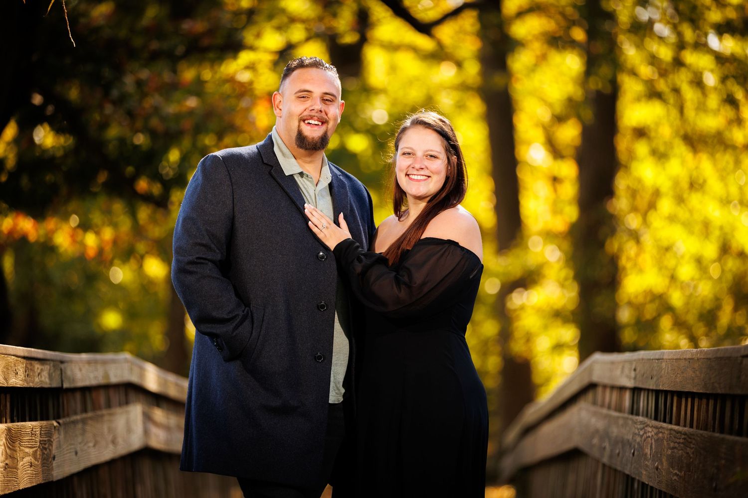 Smiling couple posing together on a wooden bridge with bright yellow autumn trees behind them.