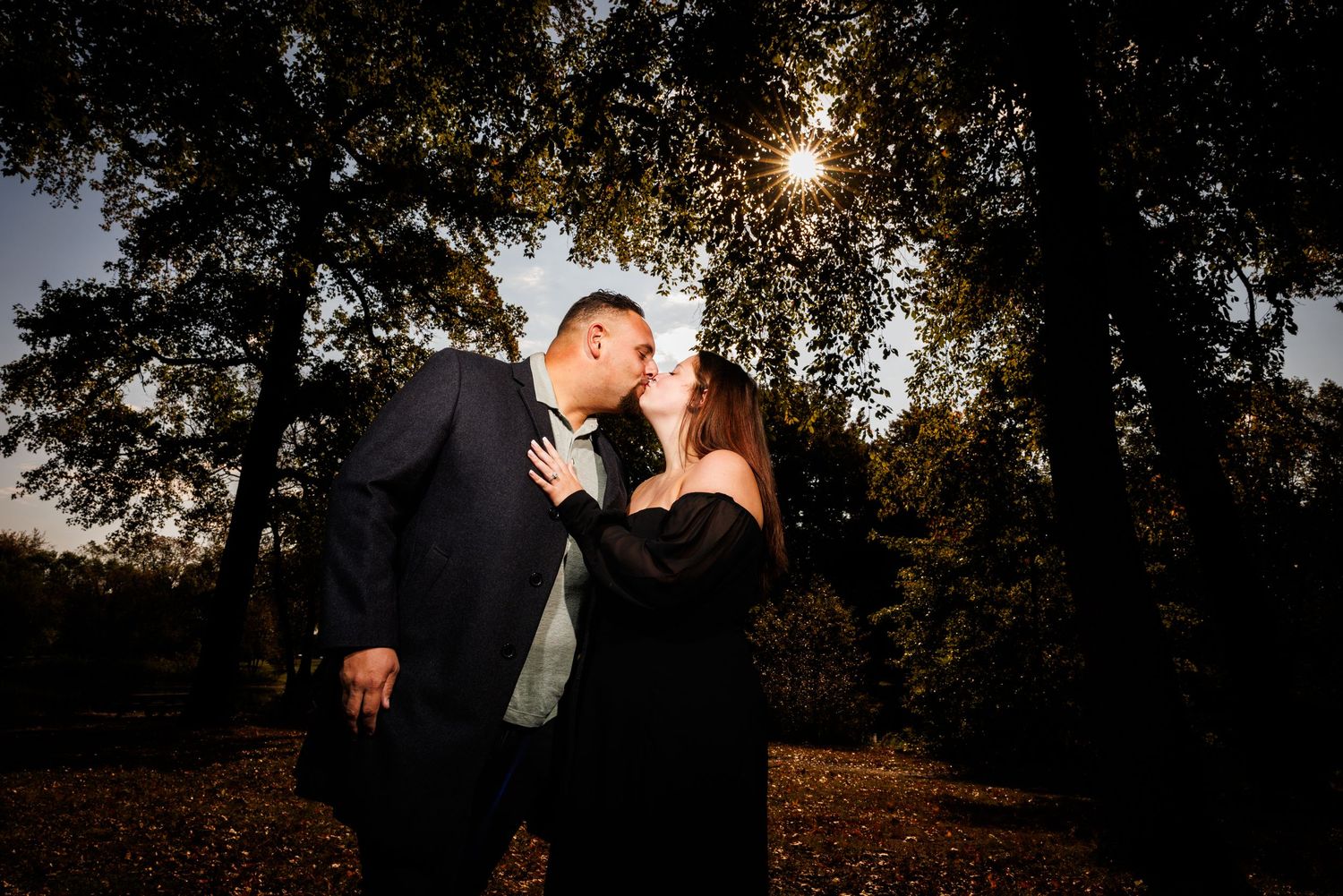 Couple sharing a tender kiss backlit by golden sunset light filtering through autumn trees.
