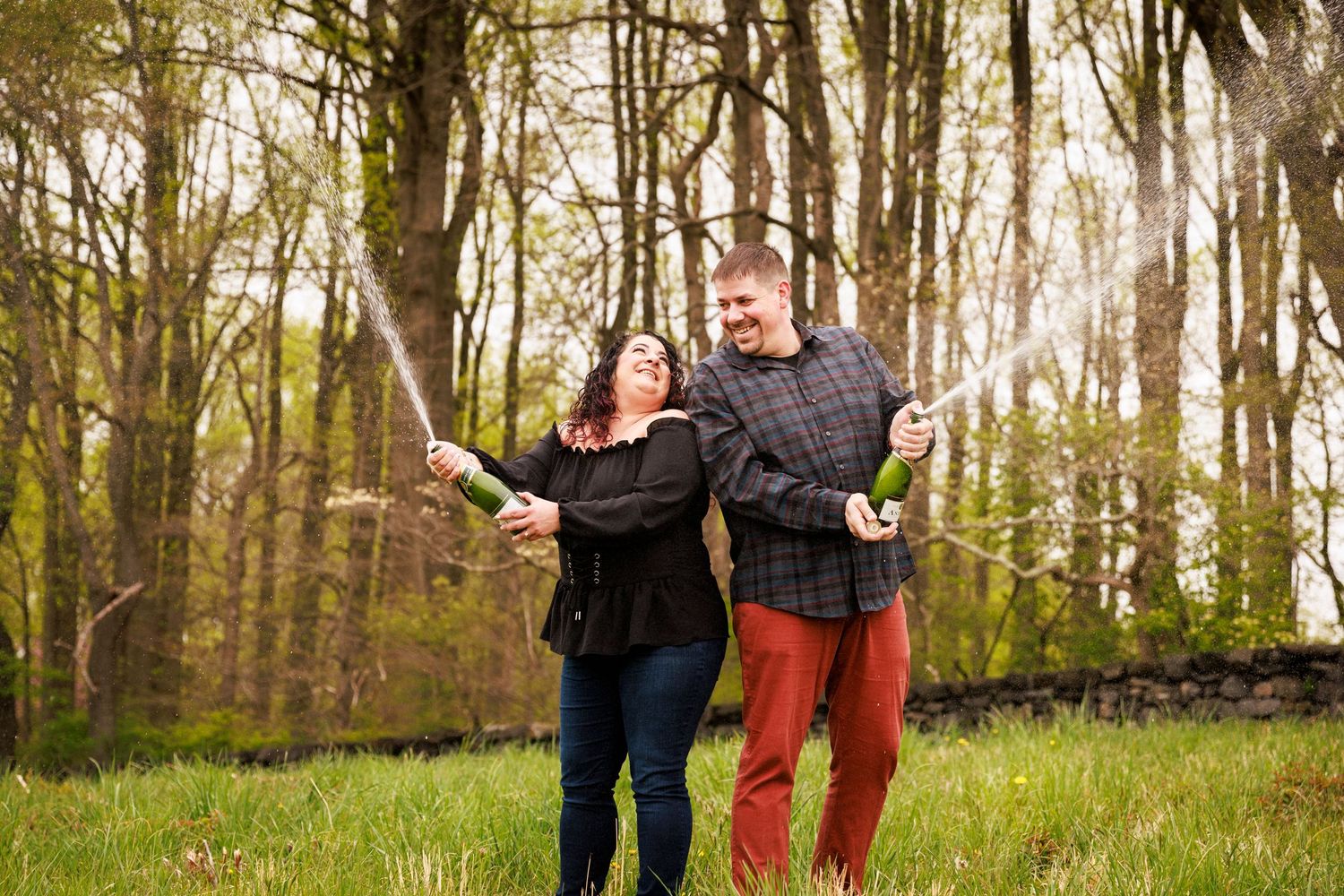 Joyful couple spraying champagne bottles in a green meadow surrounded by spring trees.