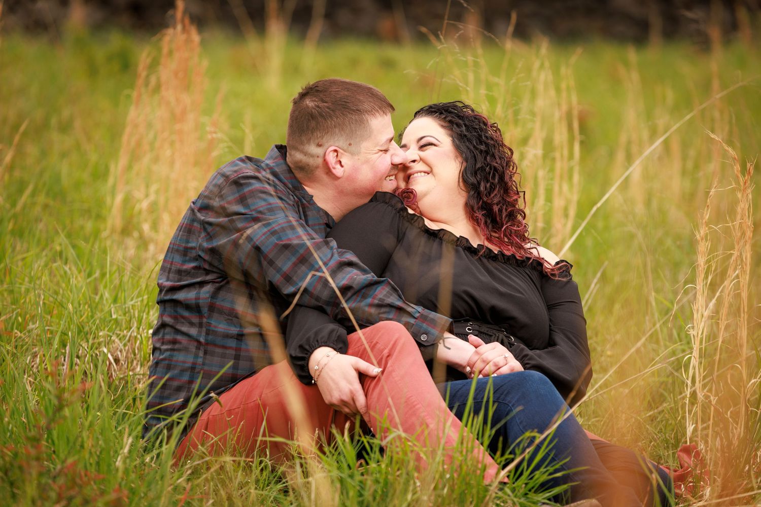 Couple sitting in tall grass field sharing a romantic moment, smiling and leaning in for a kiss.