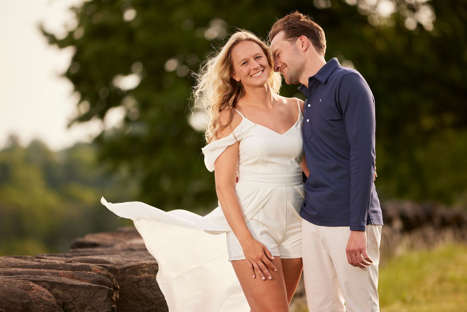 Blonde woman in white dress smiles as man in navy shirt embraces her from behind in a sunny outdoor setting.