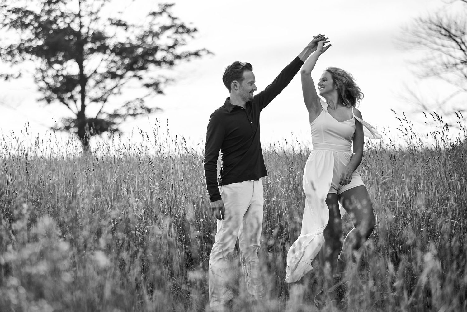 Black and white photo of a couple dancing together while holding hands in a tall grass meadow at dusk.
