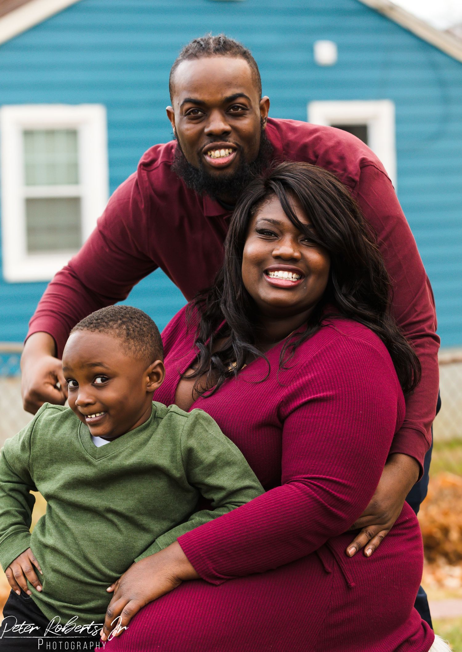 Smiling family of three, man in burgundy shirt, woman, and young boy pose together outdoors.