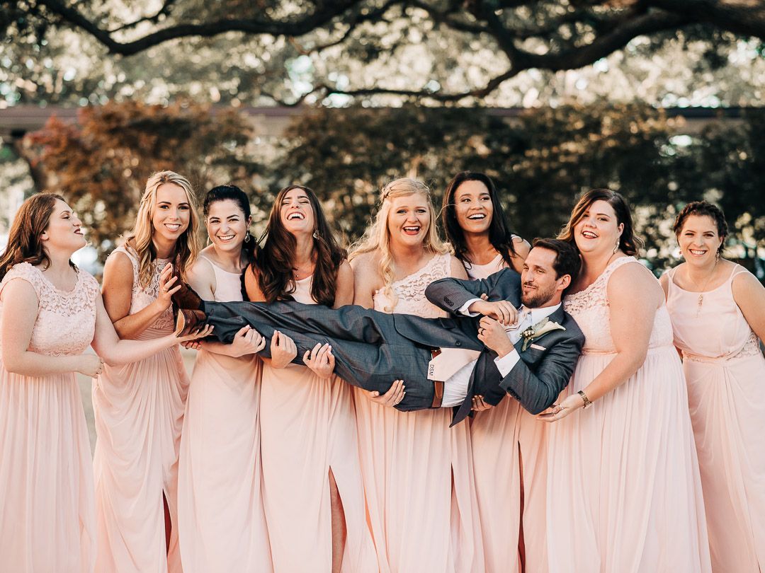 Bridesmaids in pink dresses playfully carry someone during a wedding photo session.