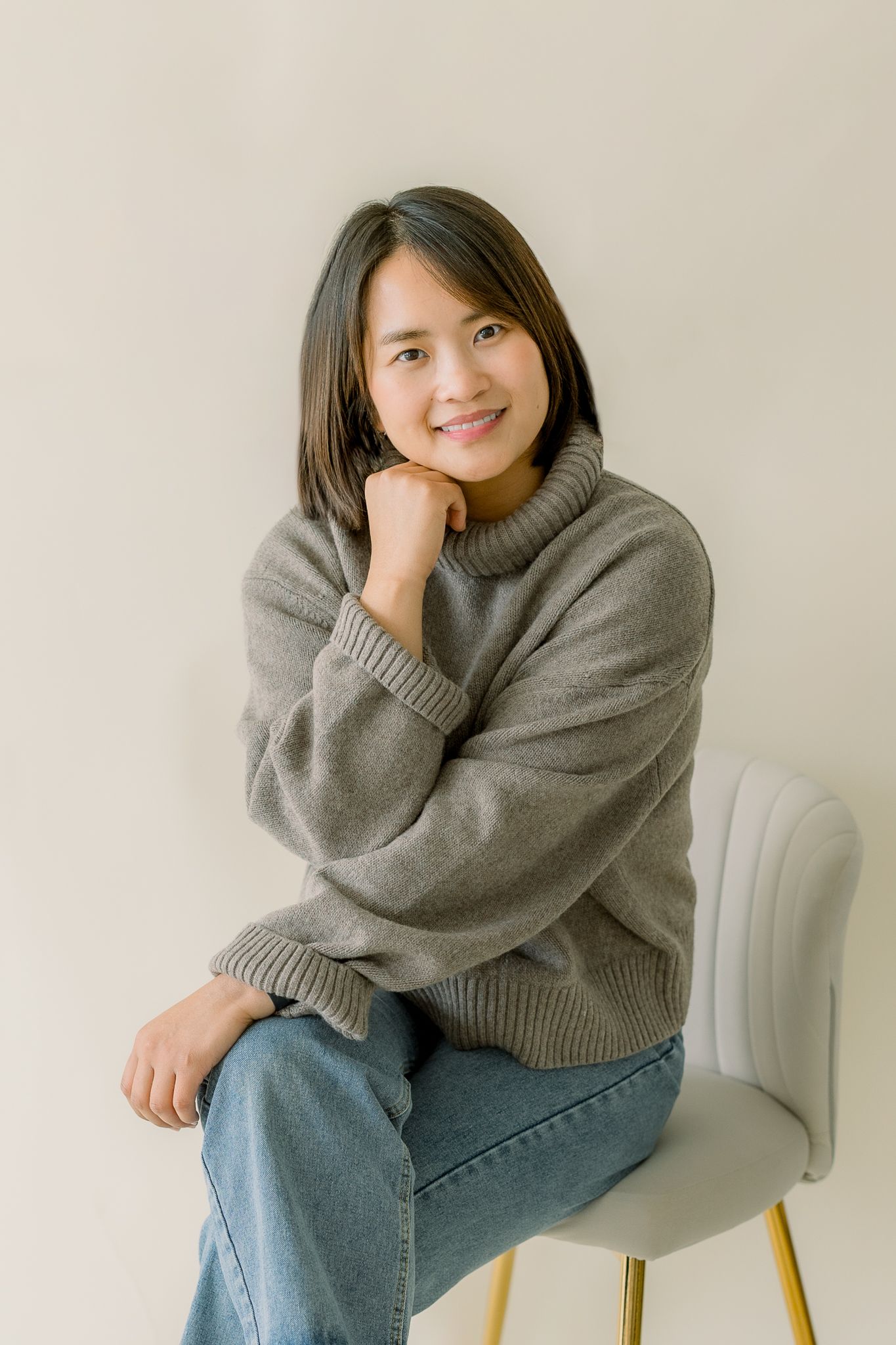 Casual lifestyle portrait of someone in a cozy gray sweater and jeans sitting on a white chair.