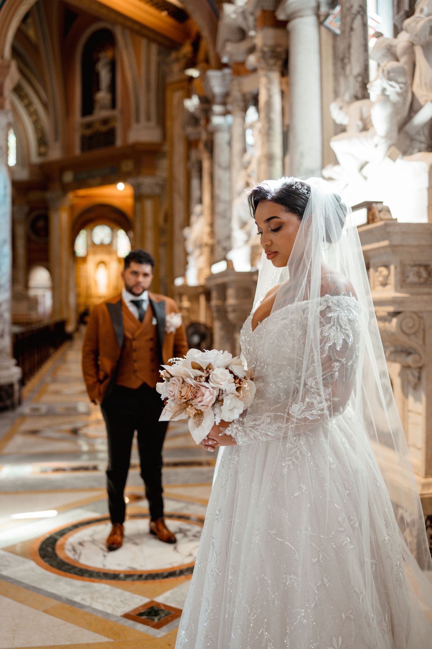 Bride in lace gown with veil holding bouquet inside ornate church with groom in background.