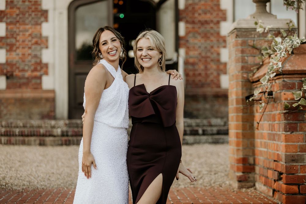 Two wedding attendees pose together outside a brick building in warm sunlight.