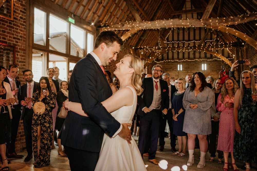 Newlyweds share first dance in rustic barn venue with fairy lights and watching guests.