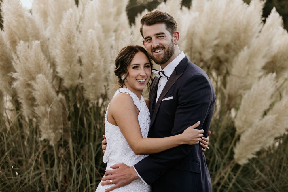 Elegant wedding couple poses together against tall decorative pampas grass in outdoor setting.