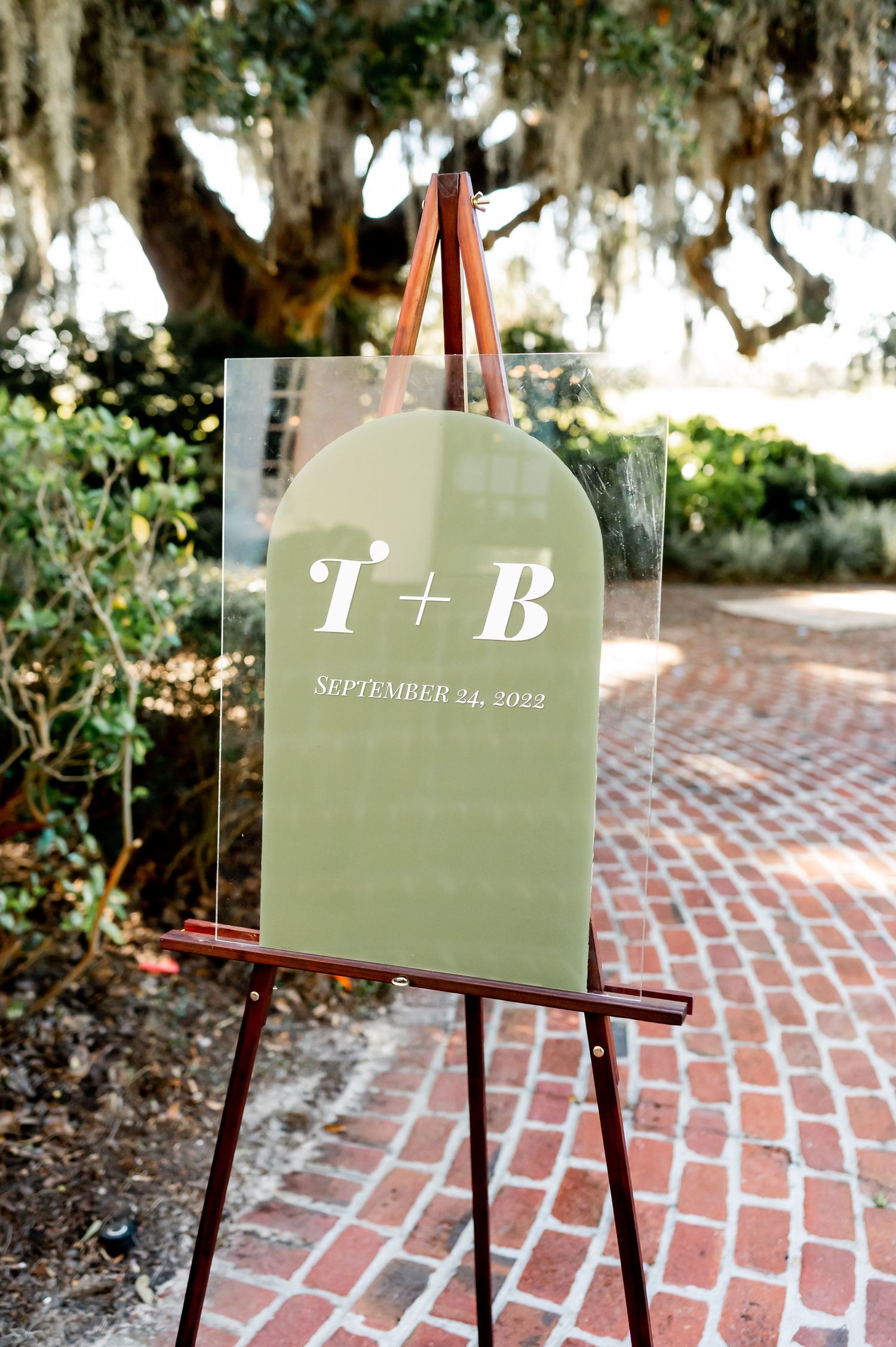 Wedding date sign displayed on easel against brick pathway with Spanish moss in background.