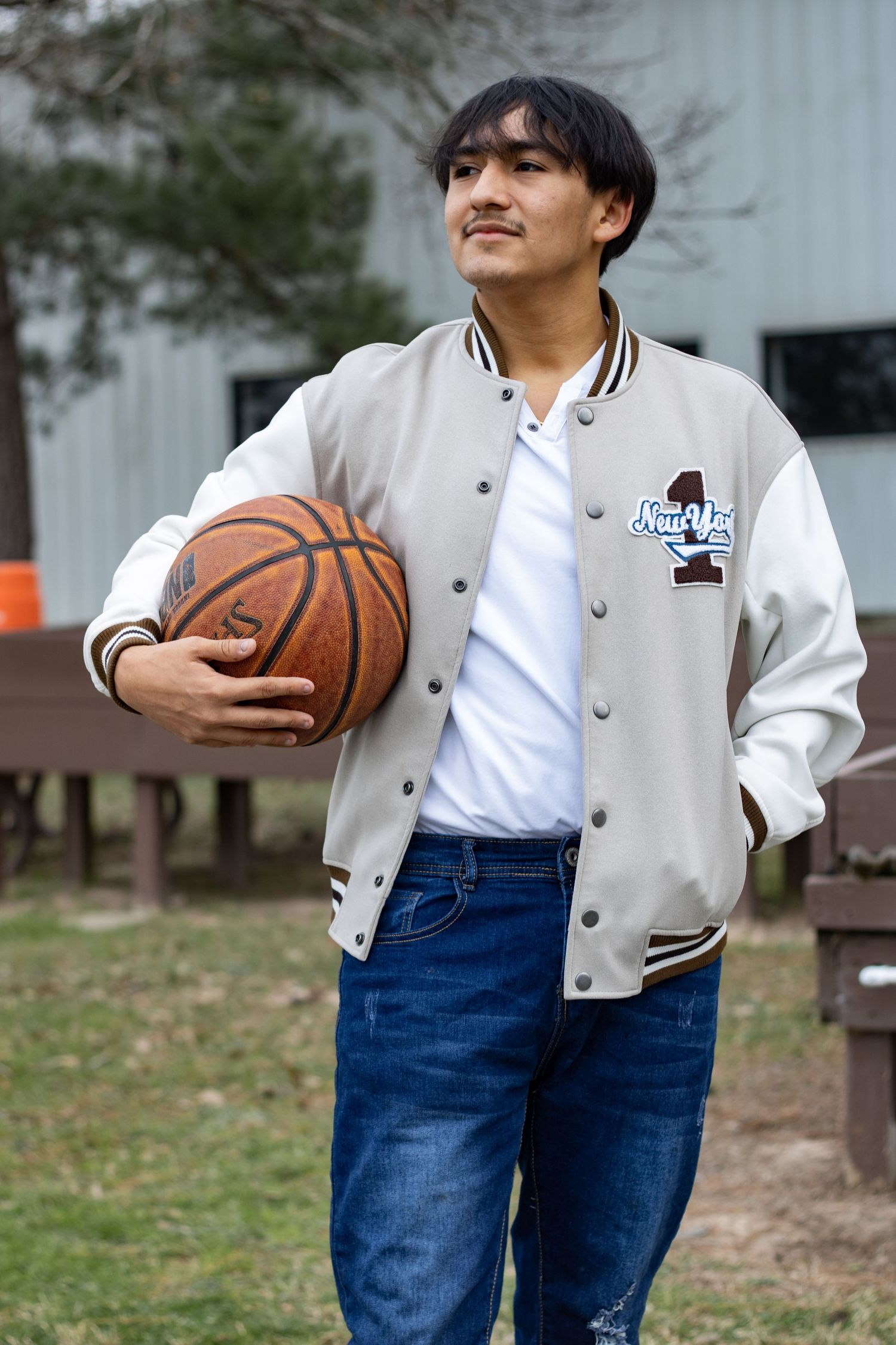 A person in a varsity jacket and jeans poses with a basketball outdoors.