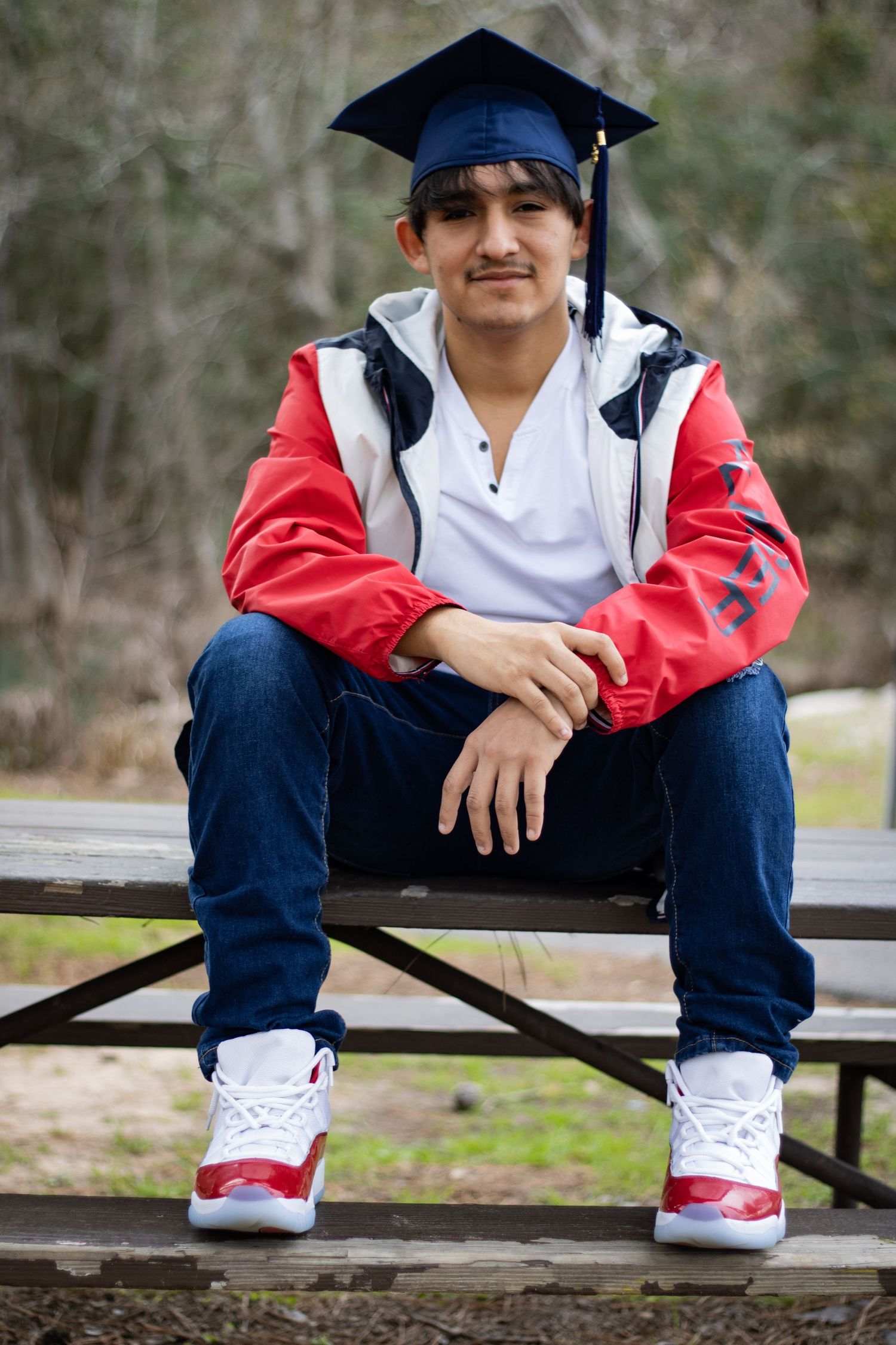 A graduate in cap and gown sits on a wooden bench wearing red and white sneakers.