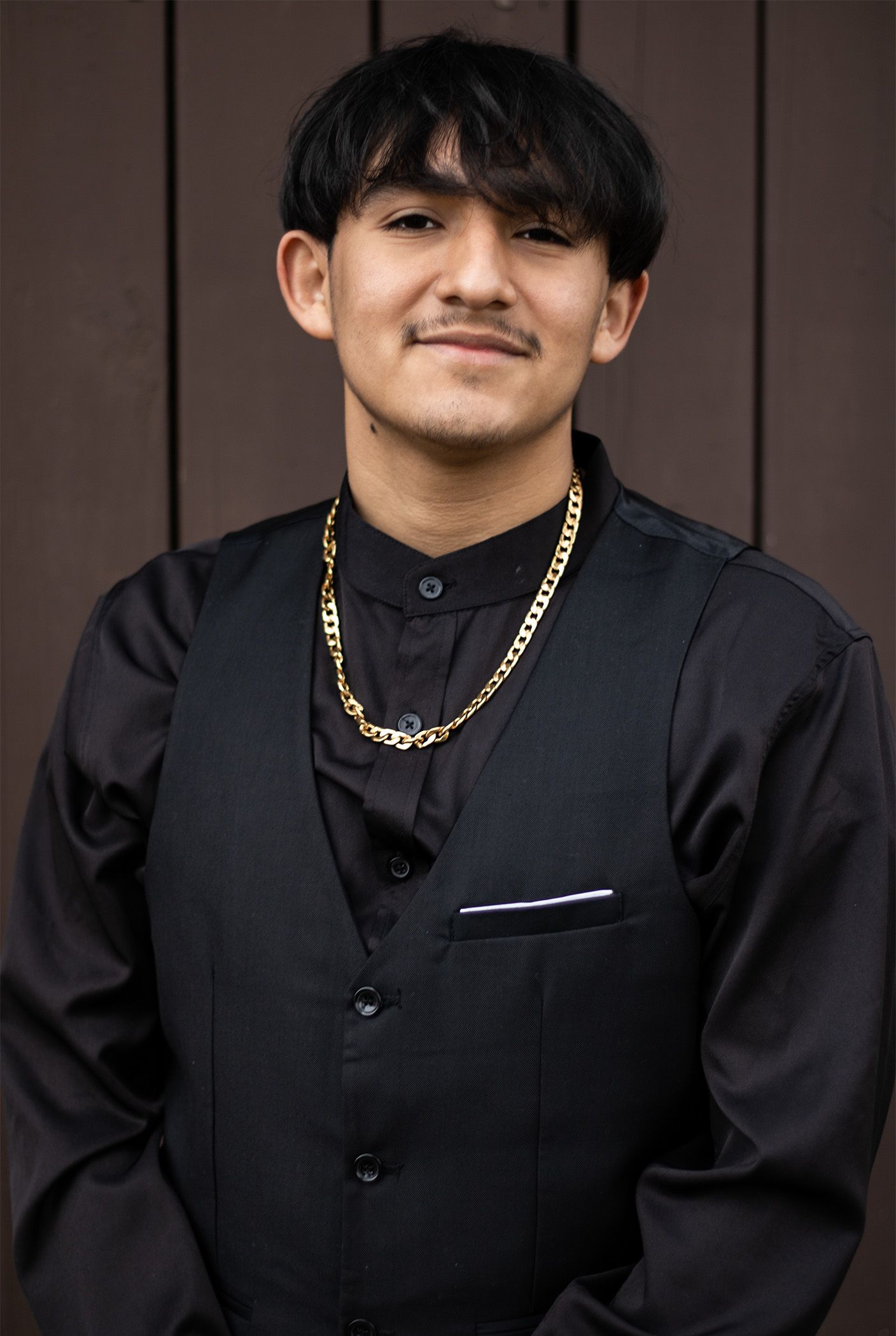 A person wearing a black button-up shirt and gold chain poses against a dark background.
