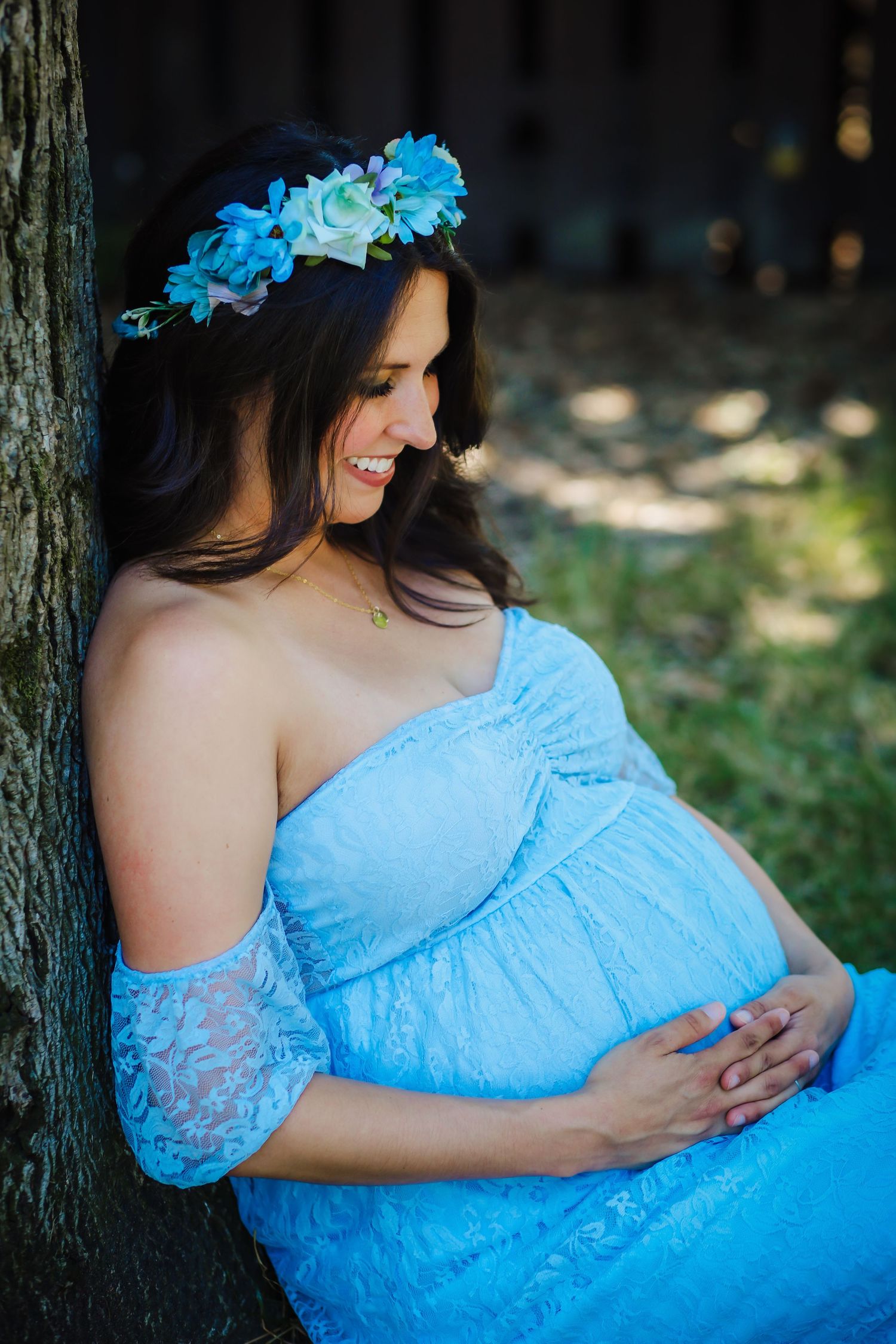 Expectant mother in flowing blue dress and floral crown poses against tree in outdoor maternity photo.