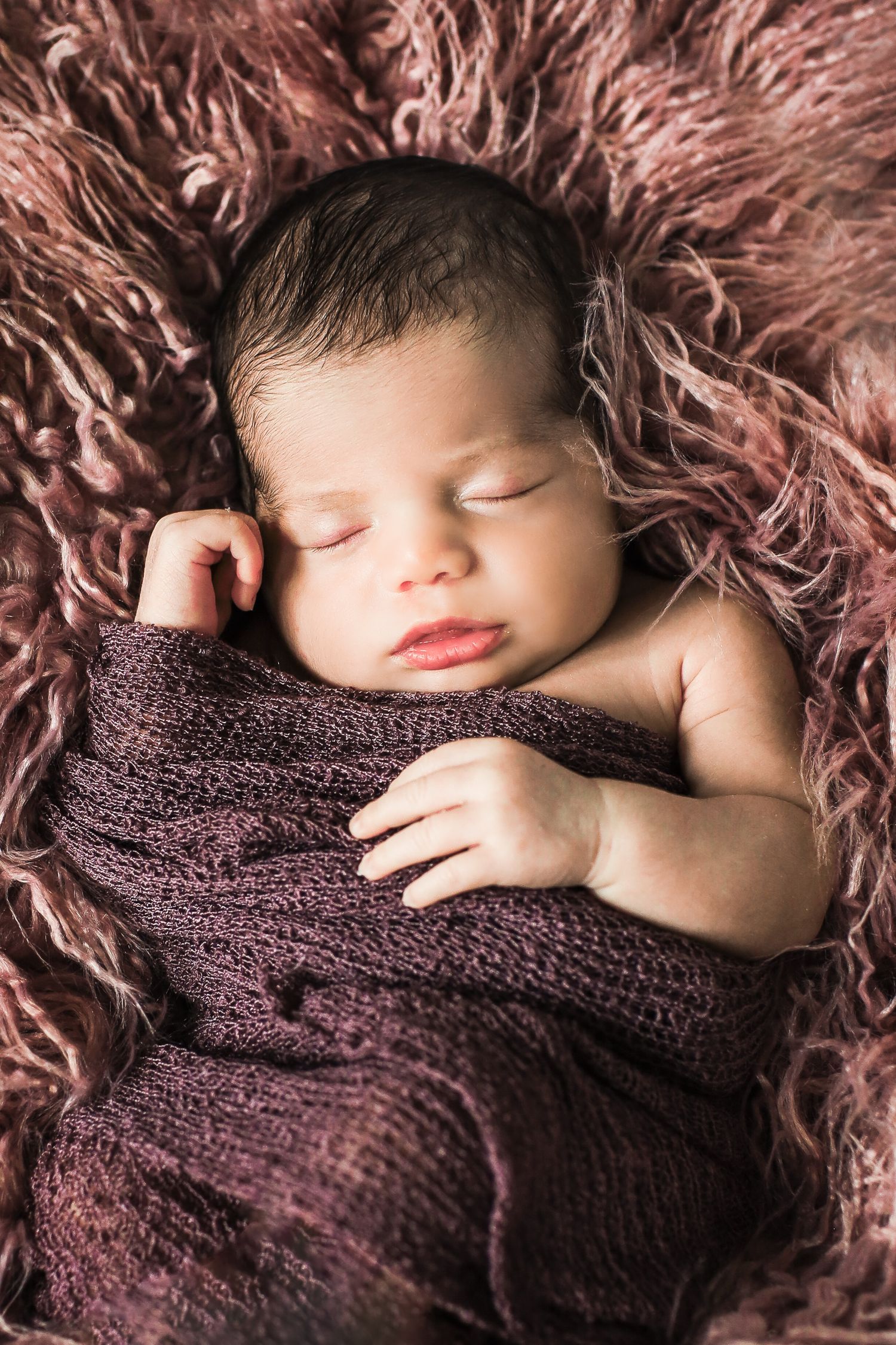 Peaceful sleeping newborn wrapped in burgundy knit blanket on textured fur backdrop.