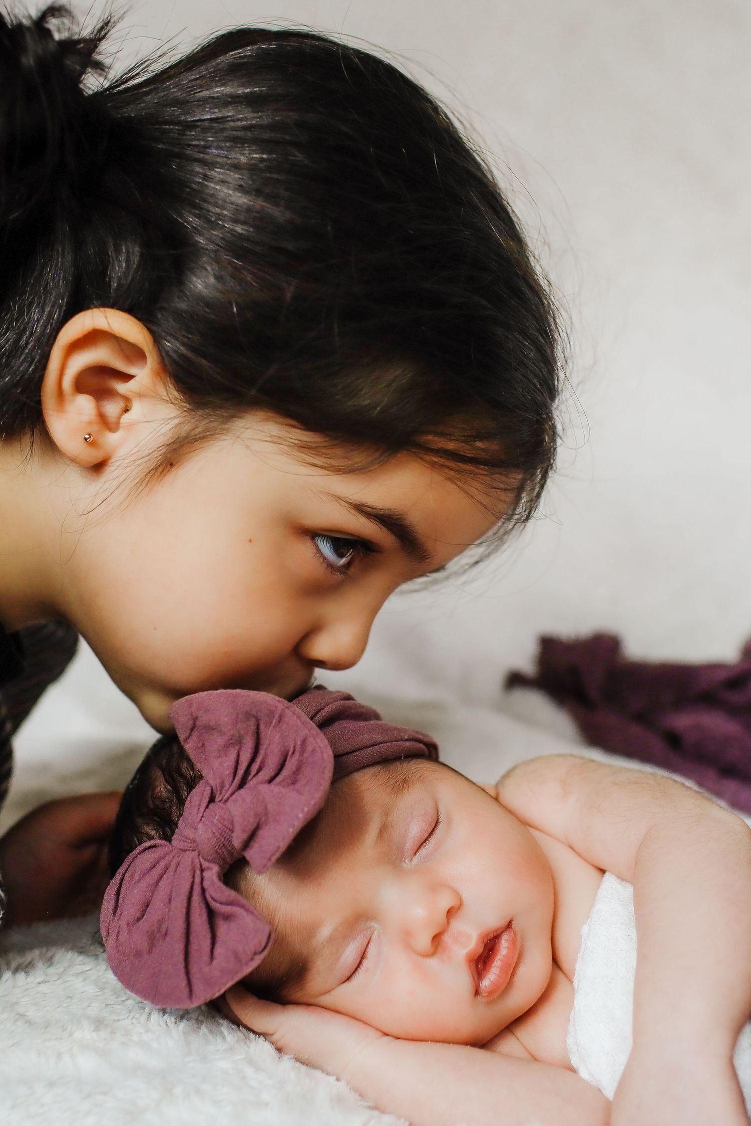 Intimate sibling bonding moment with sleeping baby wearing purple bow.