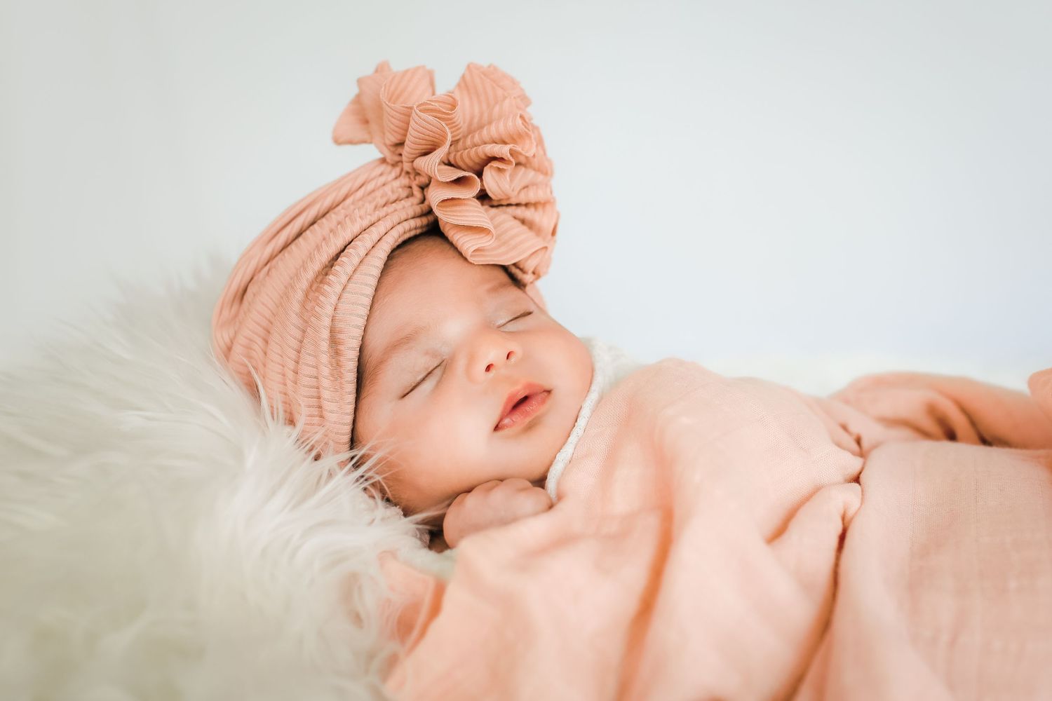 A sleeping baby wearing a peach-colored knotted turban hat and matching outfit rests on white fur bedding.
