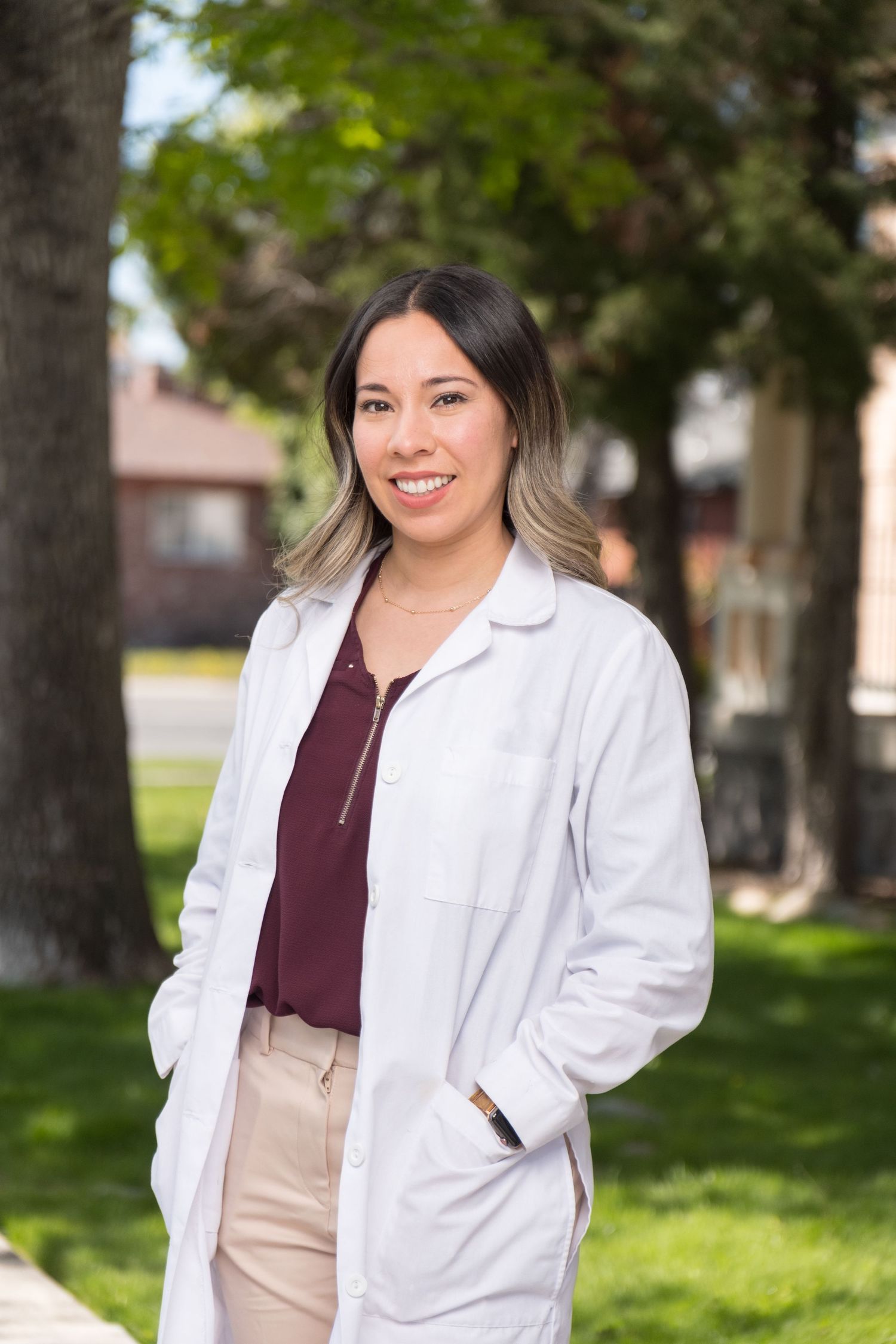 Healthcare professional in white lab coat and burgundy top stands outdoors on green lawn near trees.