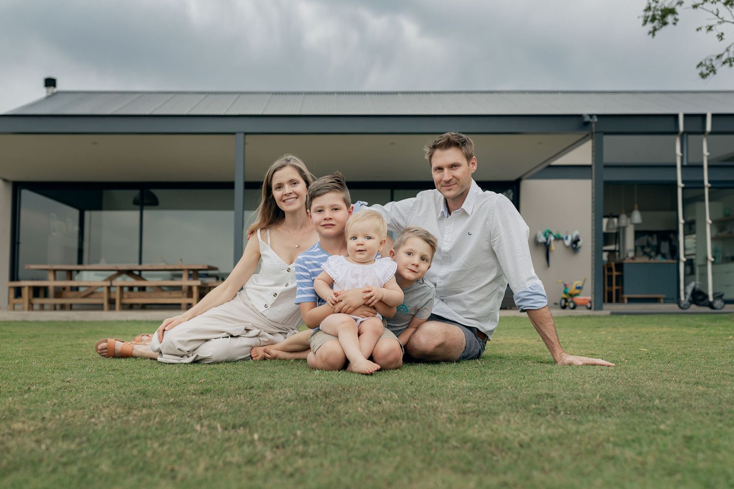 A family sits together on the lawn in front of a modern house with picnic tables.