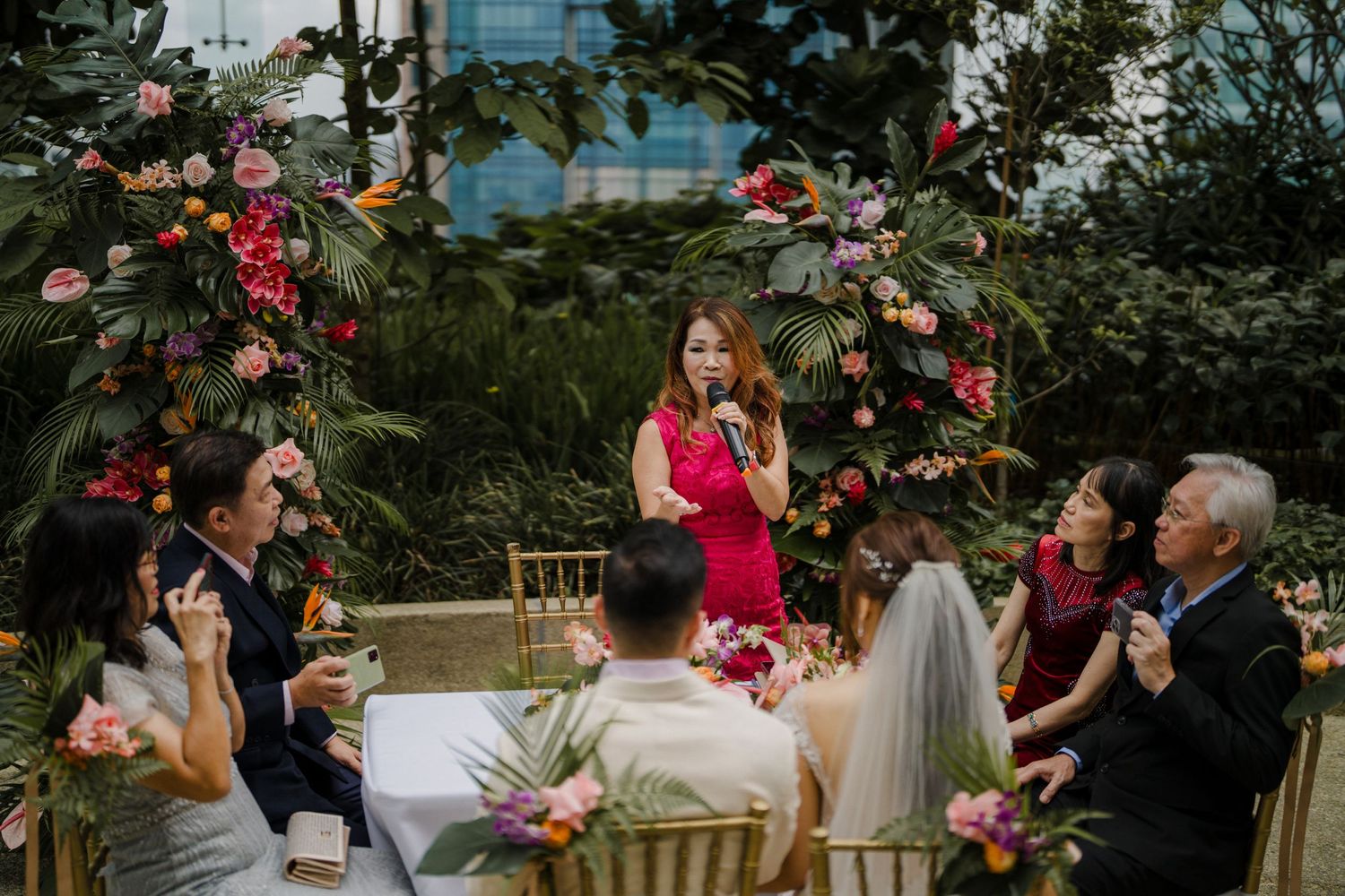Wedding ceremony guests gather outdoors surrounded by lush greenery and pink floral decorations.