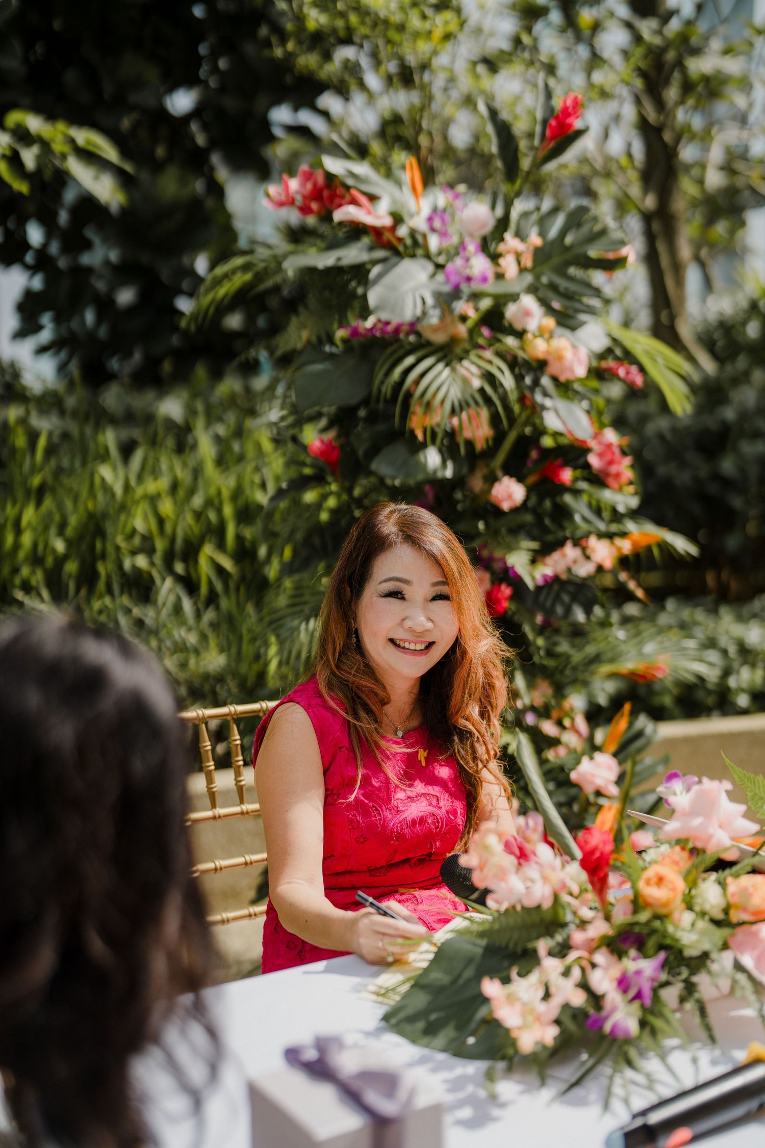 Wedding guest in red dress seated at outdoor reception with tropical floral decorations.