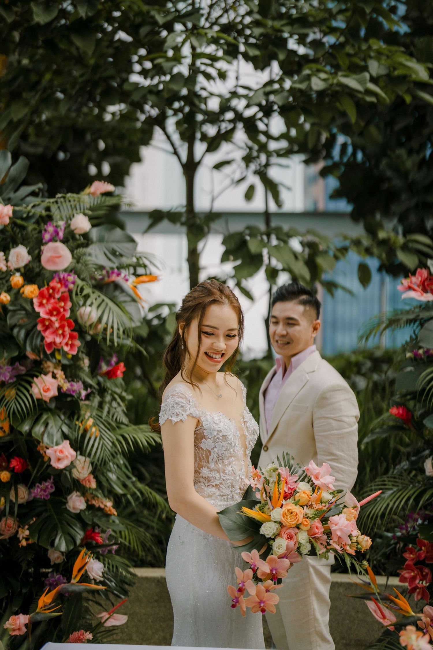A couple poses among vibrant tropical flowers and greenery in an elegant garden setting.