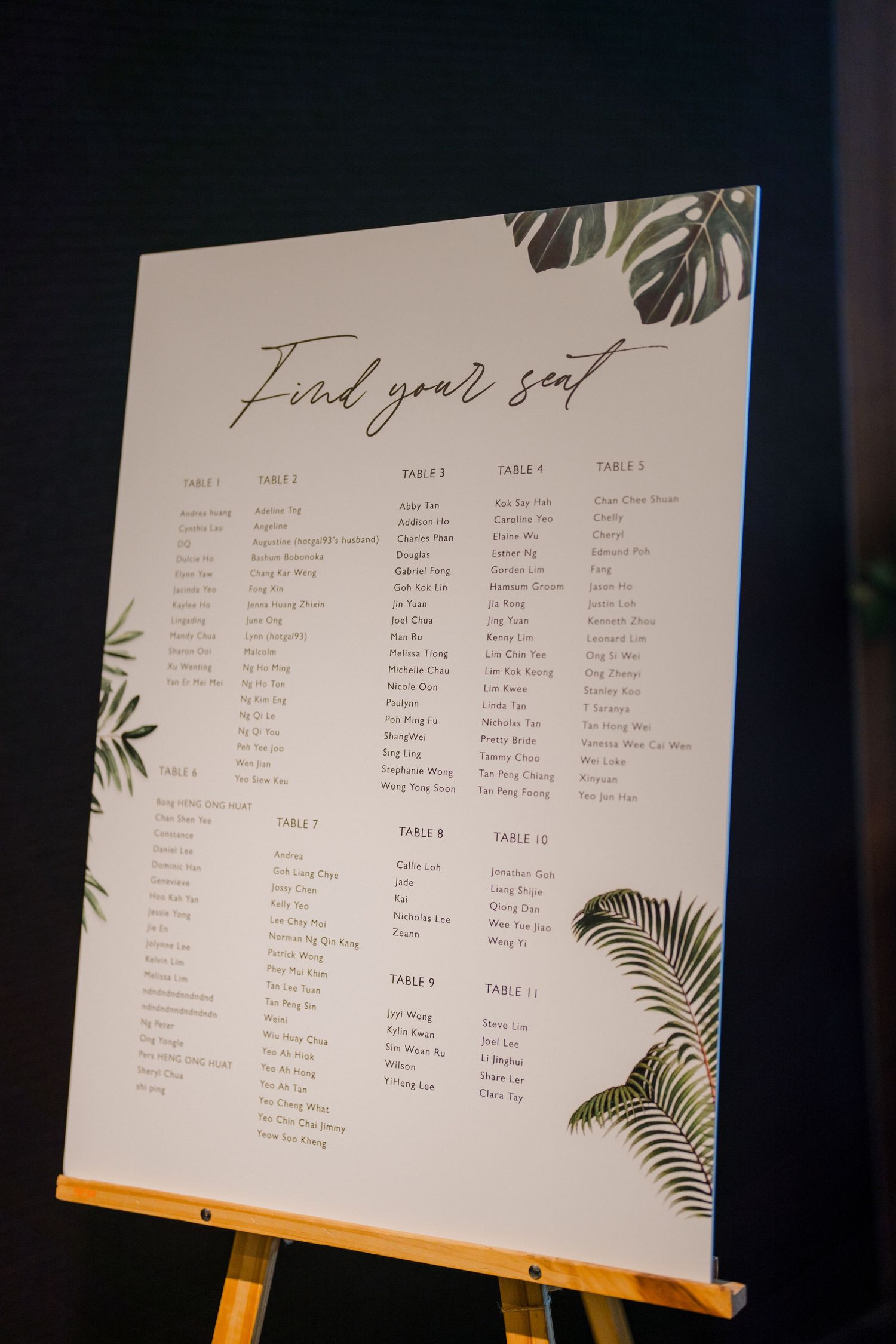 Tropical-themed wedding seating chart displayed on an easel with palm leaf decorations.