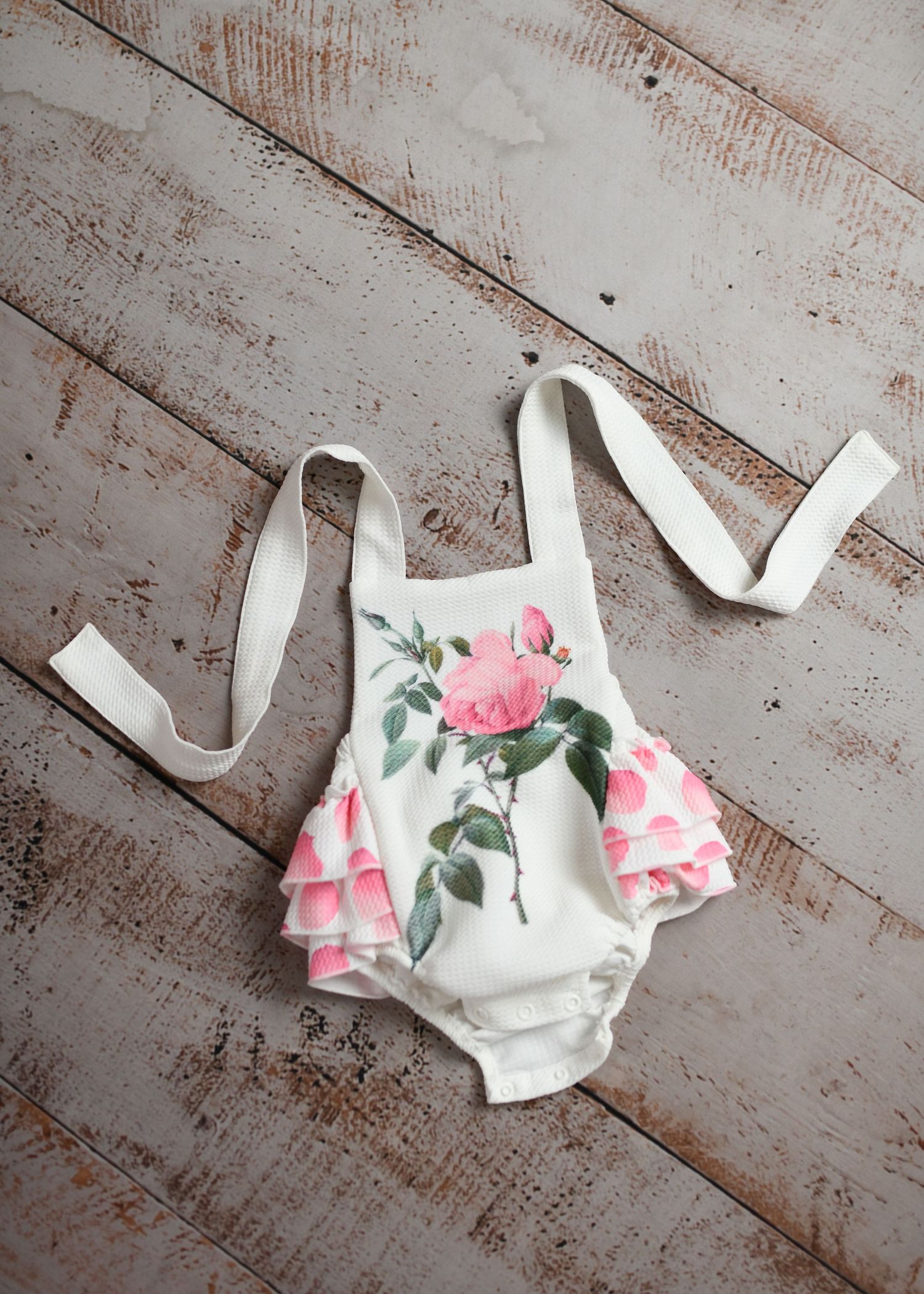 White floral romper with pink rose pattern laid flat on rustic wooden surface.