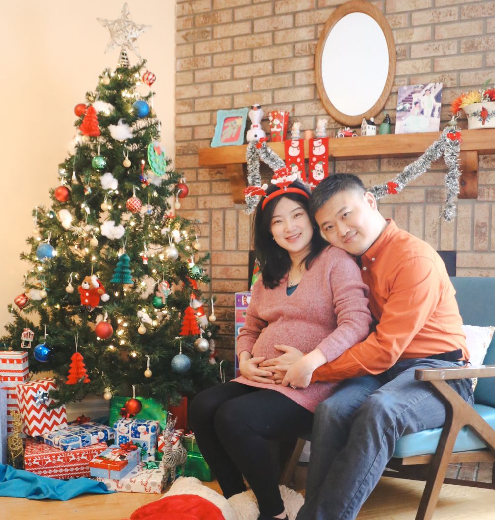 Couple posing by decorated Christmas tree with wrapped presents underneath.