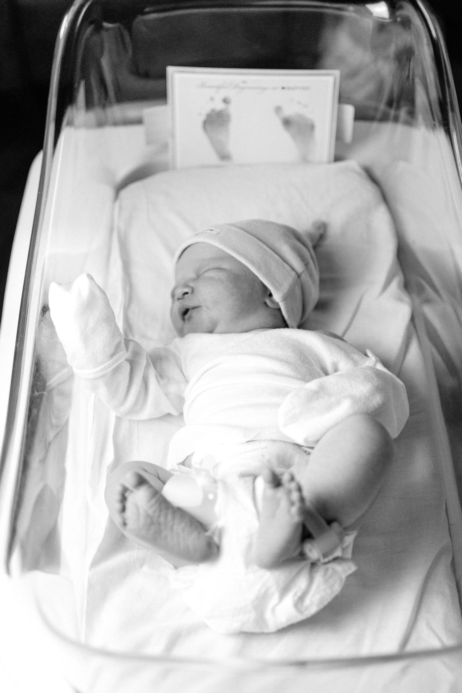 Black and white photo of newborn baby lying in hospital bassinet with footprint card in background.