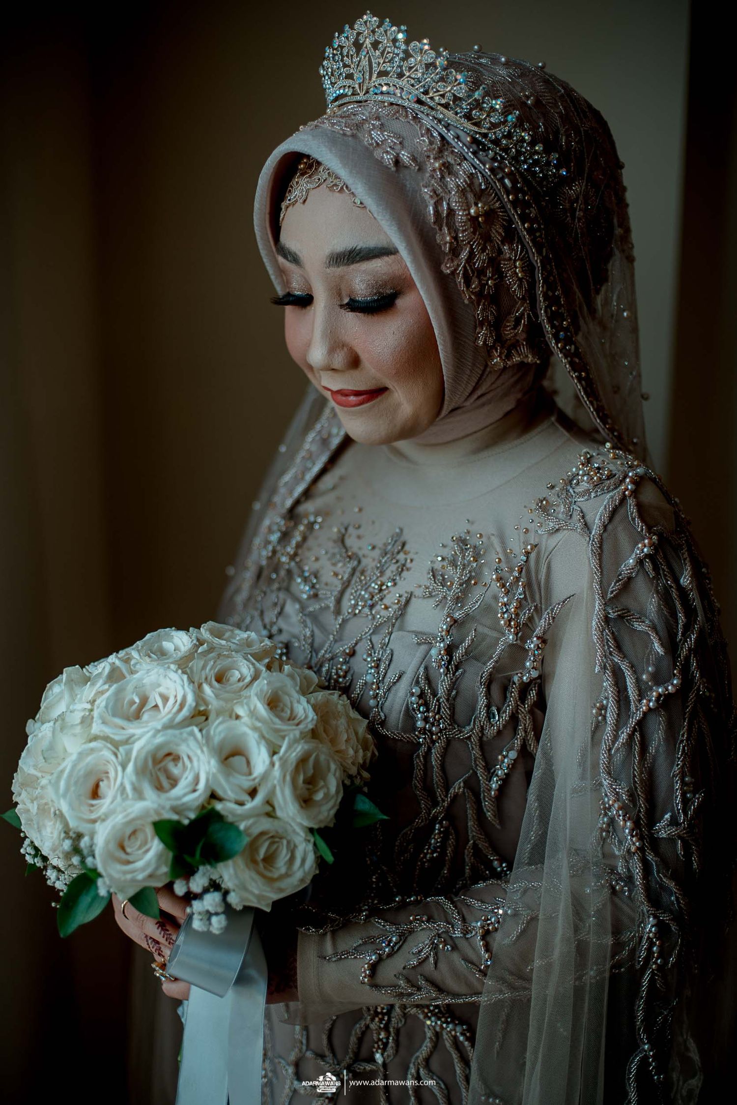 A bride in an ornate hijab and beaded gown holds a white rose bouquet.