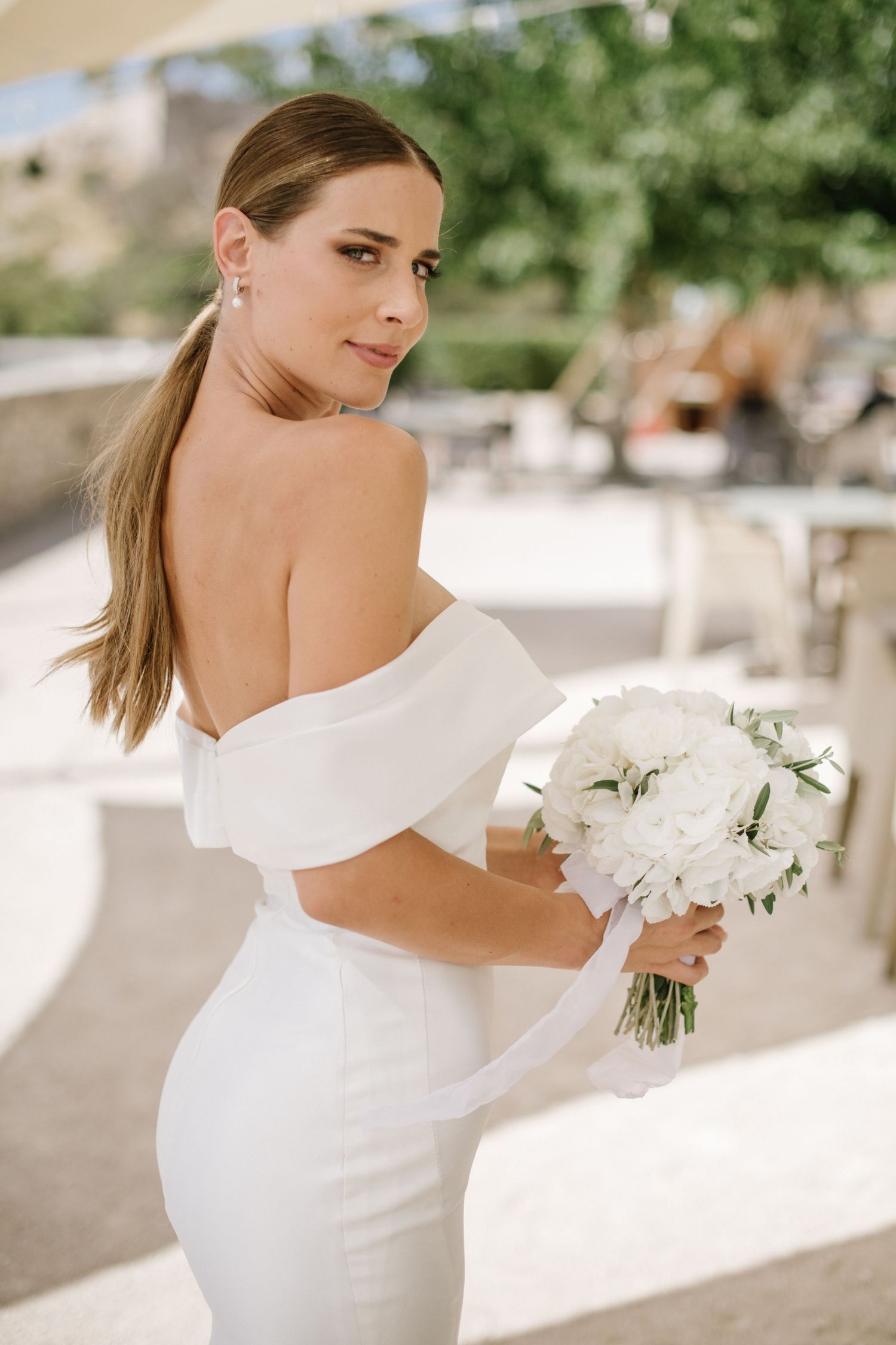 Beautiful bride in off-shoulder white wedding dress holds elegant white floral bouquet while looking over her shoulder.