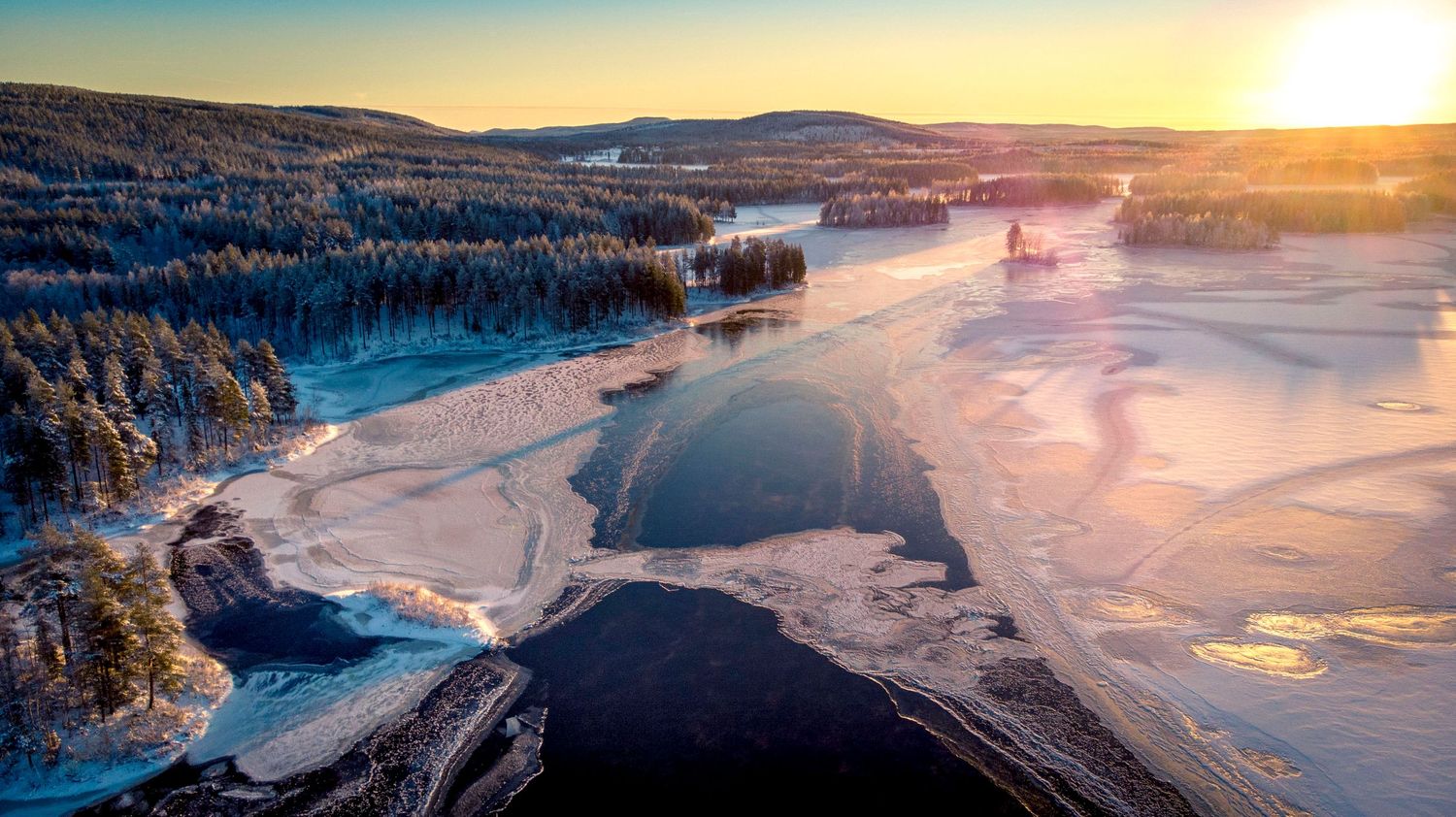 Aerial sunset view of a partially frozen lake surrounded by snow-covered forests in winter.
