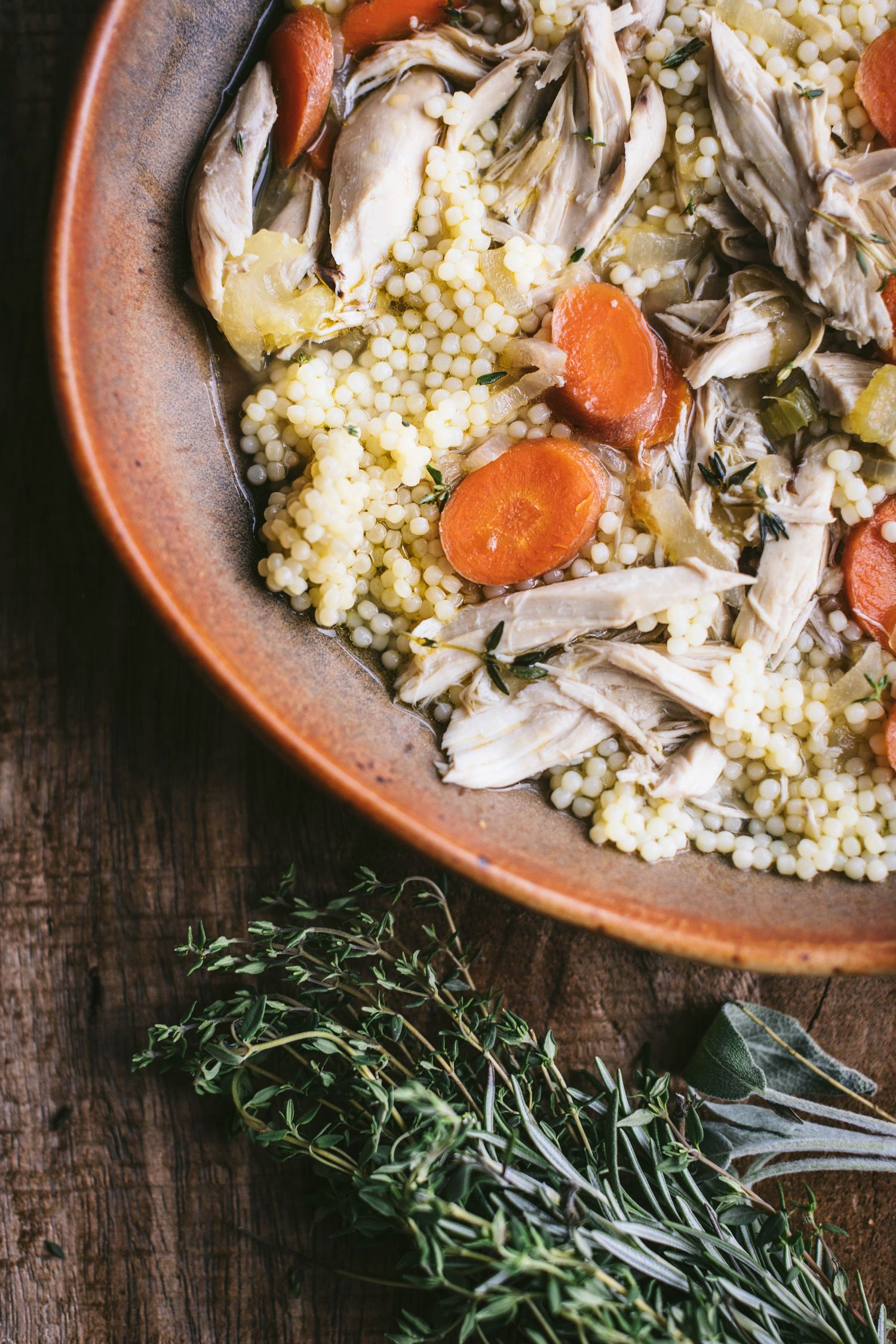 Close-up of shredded chicken with pearl couscous and carrots in a clay bowl, garnished with fresh thyme and sage.
