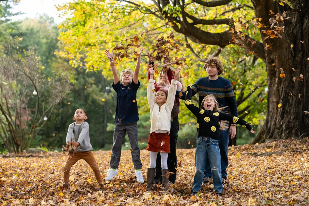 Group of children playing and jumping in autumn leaves under yellow trees.