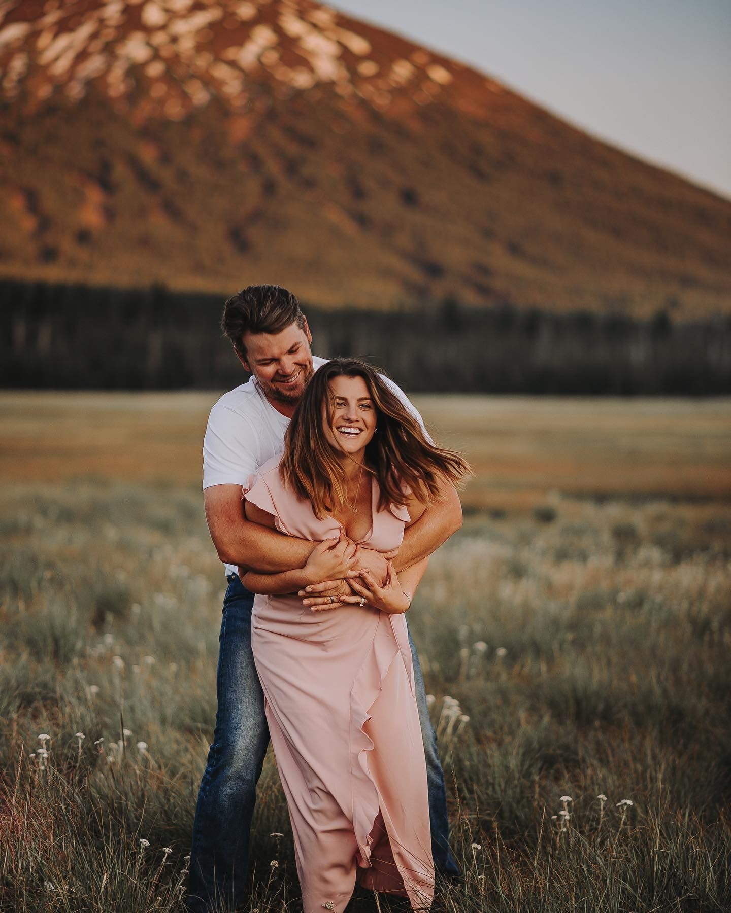 Couple laughing and embracing in a field at sunset with mountains in the background.