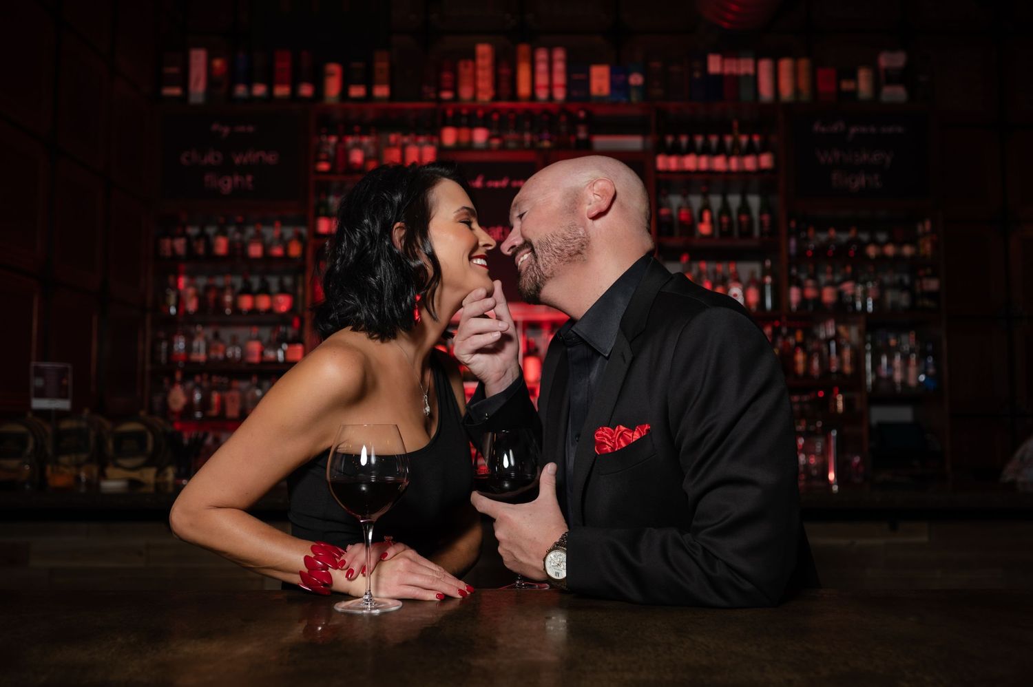 Romantic couple sharing wine and laughter at dimly lit bar with bottles in background.