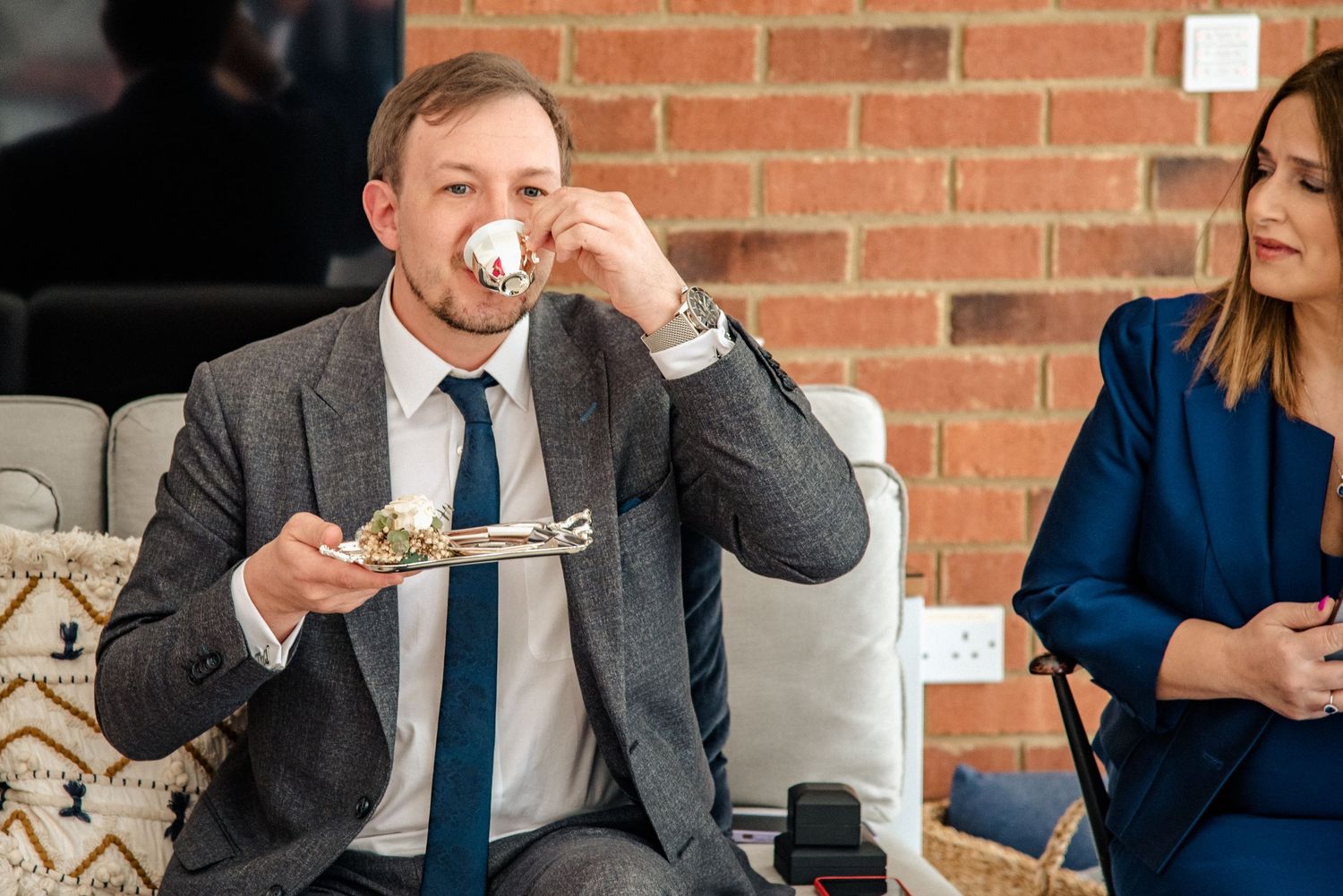 A person in a grey suit and tie enjoying food and coffee at a cafe with a brick wall backdrop.