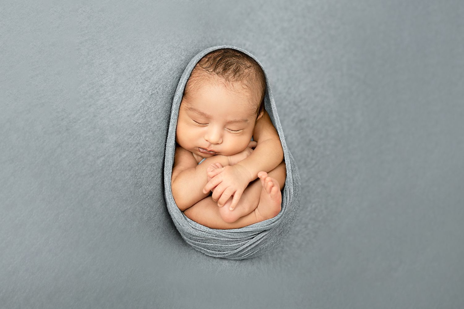 Artistic newborn photography with baby wrapped in soft grey fabric against grey background.