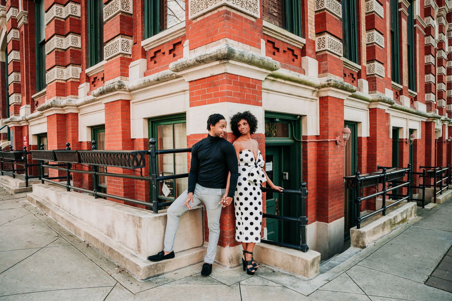 Two people in casual attire pose together at the corner entrance of a red brick vintage building with ornate architectural details.
