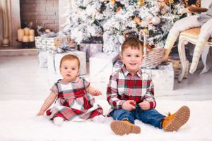 Two children in coordinating plaid outfits sitting in front of a decorated Christmas tree with presents.