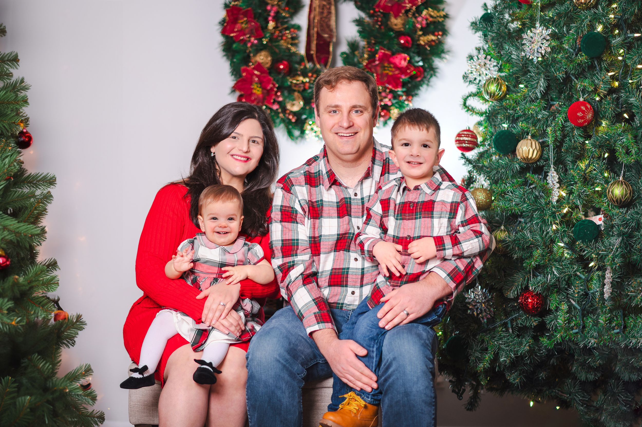 Holiday family photo featuring coordinated red and plaid outfits by decorated Christmas trees.