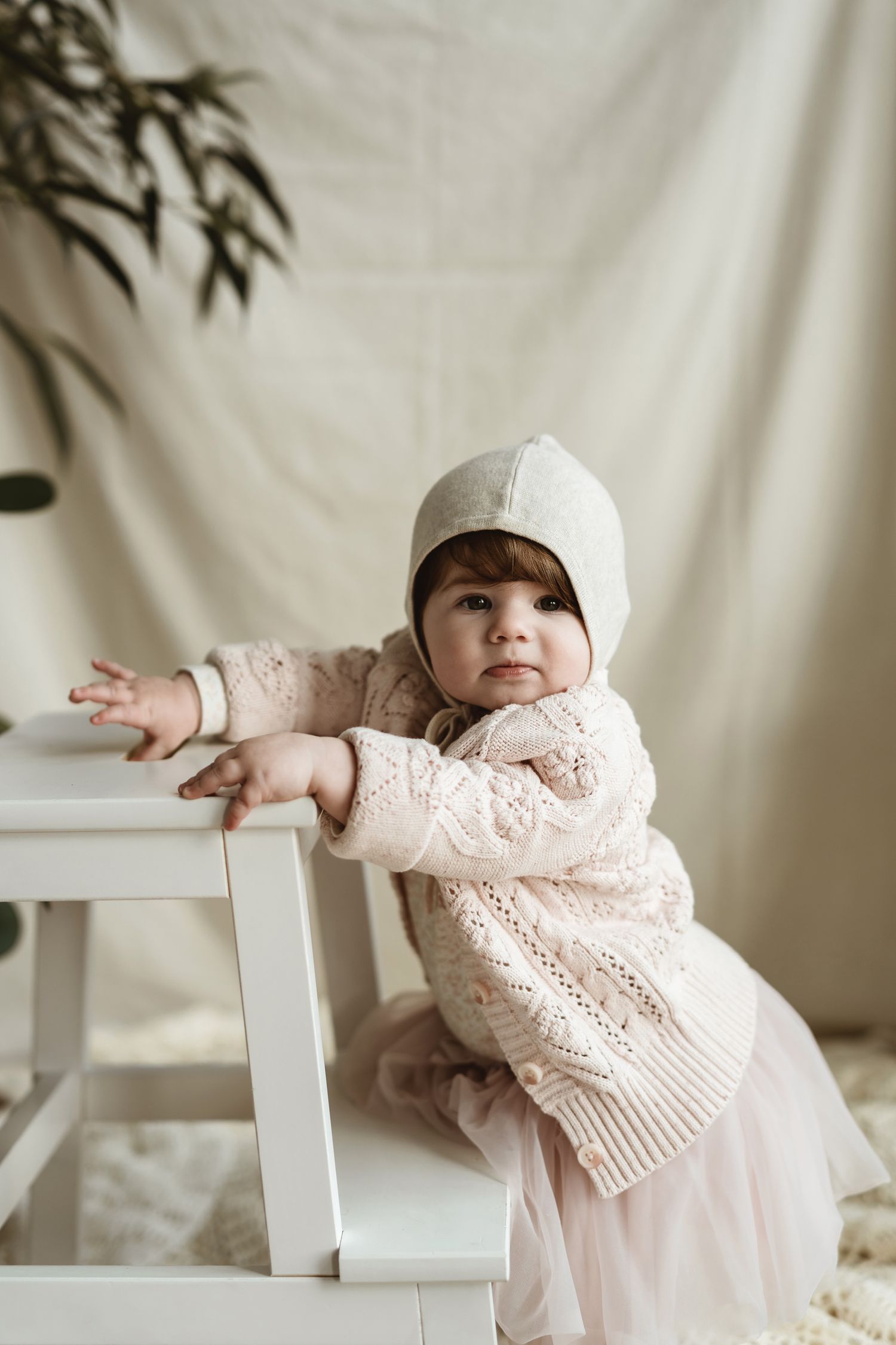Toddler in white bonnet and lace dress playing with white chair against cream backdrop.