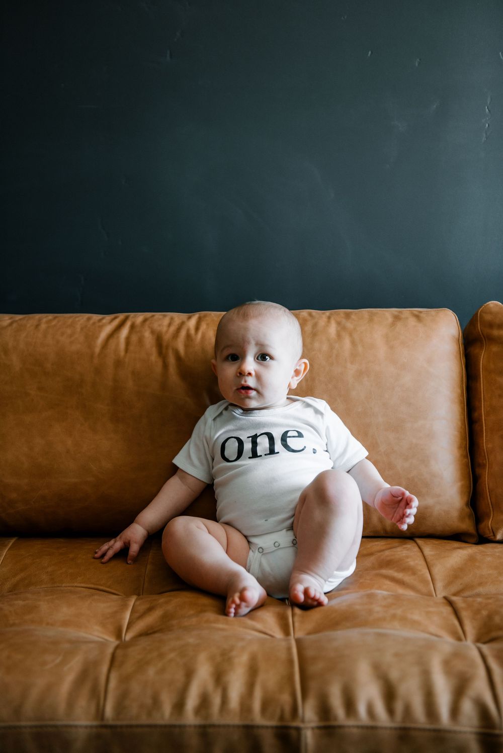 An infant in white clothing with text sits on a tan leather sofa.
