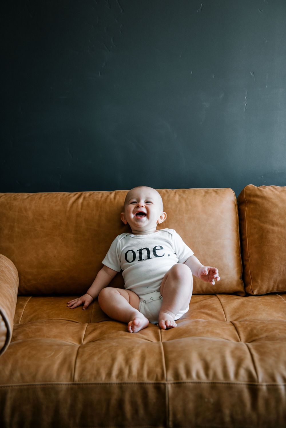 A baby wearing a 'one' onesie sits on a brown leather couch.