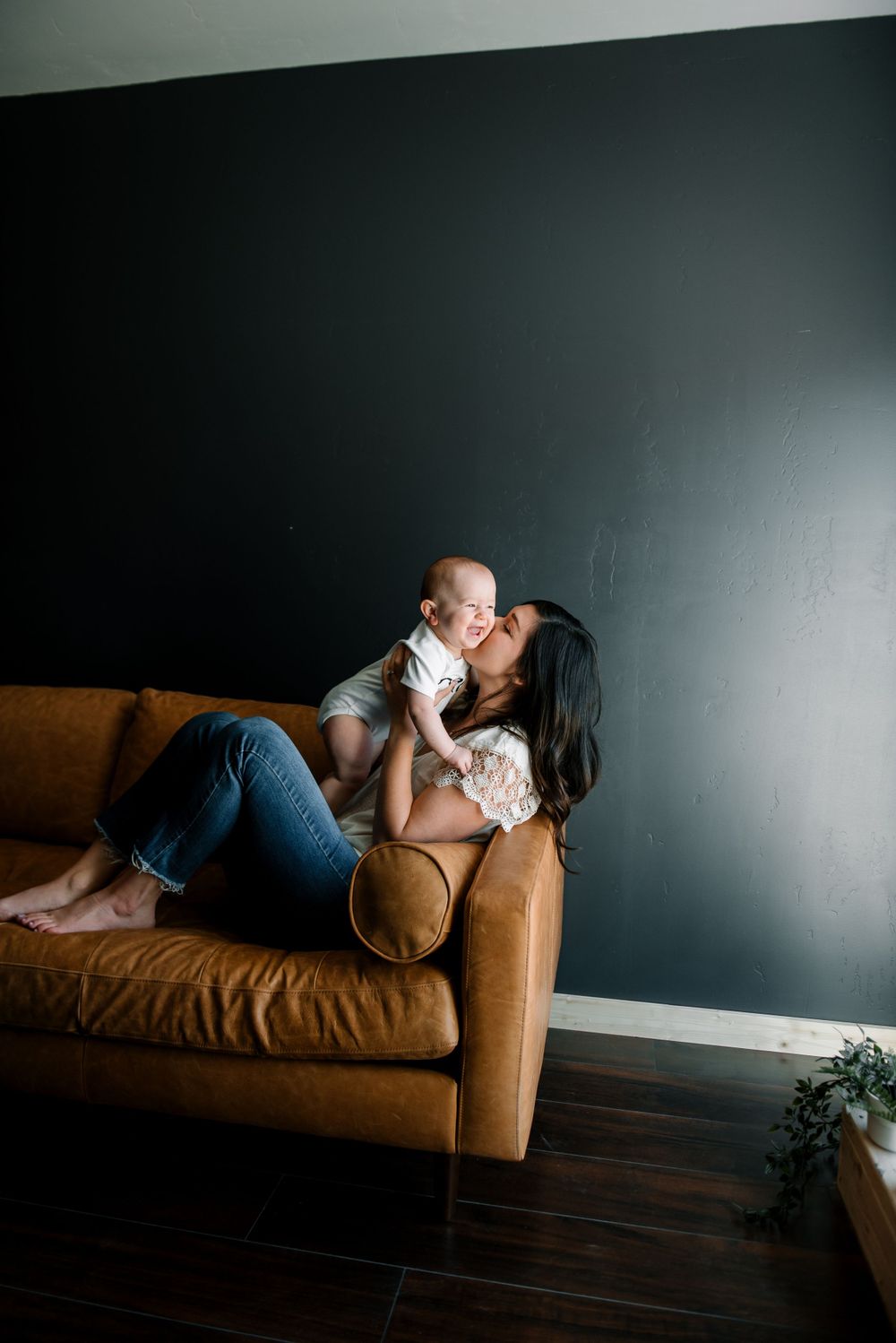 Mother and baby share tender moment on brown leather couch against dark wall.